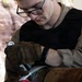 A young male technician wearing glasses and Mechanix brand tactical gloves is working on a large cylindrical metallic object, using red-handled pliers or cutters to remove or cut a fabric/tape band. The individual is dressed in military-style olive/tan clothing and a black undershirt, working outdoors in what appears to be a field environment. Stenciled markings are visible on the cylindrical object.