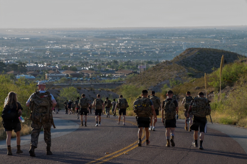 A large group of individuals wearing camouflage gear and carrying heavy backpacks (rucksacks) are walking along a paved two-lane road descending from an elevated position. The terrain appears arid and semi-desert, with scrub vegetation and hills visible. A sprawling urban area is visible in the valley below, suggesting this is a military-style ruck march or fitness event.