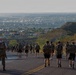 A large group of individuals wearing camouflage gear and carrying heavy backpacks (rucksacks) are walking along a paved two-lane road descending from an elevated position. The terrain appears arid and semi-desert, with scrub vegetation and hills visible. A sprawling urban area is visible in the valley below, suggesting this is a military-style ruck march or fitness event.