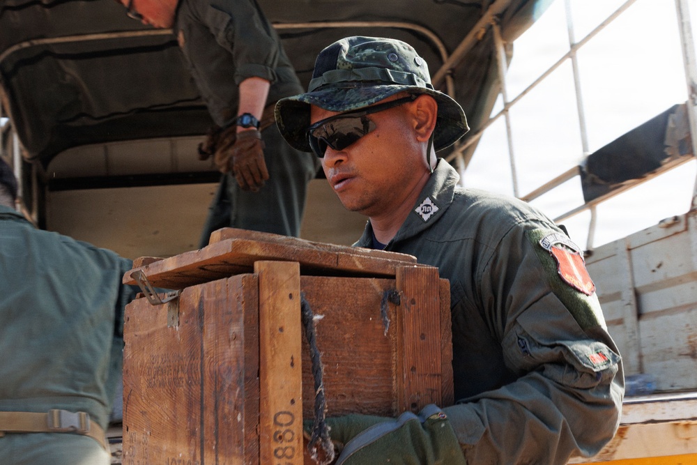 A military personnel wearing olive green uniform, boonie hat, and sunglasses is handling a wooden crate near what appears to be a military vehicle or truck. The individual has visible patches on their uniform sleeve, including what appears to be a flag patch. At least one other uniformed person is visible in the background.