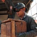 A military personnel wearing olive green uniform, boonie hat, and sunglasses is handling a wooden crate near what appears to be a military vehicle or truck. The individual has visible patches on their uniform sleeve, including what appears to be a flag patch. At least one other uniformed person is visible in the background.