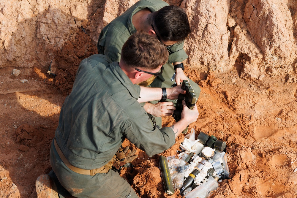 Two individuals wearing olive/sage green military-style uniforms are working in a shallow excavated pit with red clay soil. They appear to be handling and examining ordnance or explosive-related items collected in the pit. The activity is consistent with Explosive Ordnance Disposal (EOD) or demining operations.
