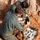 U.S. Marines and Thailand Mine Action Center participate in Deployed for Training Bilateral EOD Exercise Two individuals wearing olive/sage green military-style uniforms are working in a shallow excavated pit with red clay soil. They appear to be handling and examining ordnance or explosive-related items collected in the pit. The activity is consistent with Explosive Ordnance Disposal (EOD) or demining operations.