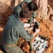 Two individuals wearing olive/sage green military-style uniforms are working in a shallow excavated pit with red clay soil. They appear to be handling and examining ordnance or explosive-related items collected in the pit. The activity is consistent with Explosive Ordnance Disposal (EOD) or demining operations.