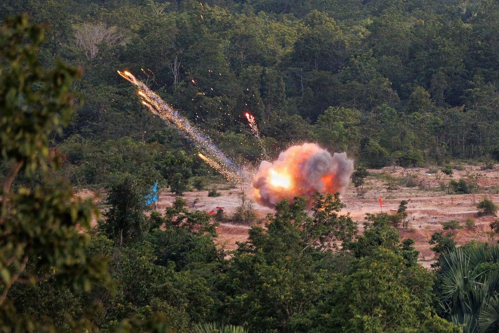 The image captures a large explosion with an orange fireball and smoke cloud occurring in an open sandy area surrounded by dense tropical forest. Bright projectile trails with sparks are visible arcing upward and outward from the blast point. Colored marker flags are visible in the field, suggesting a controlled or designated detonation zone.