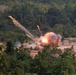 The image captures a large explosion with an orange fireball and smoke cloud occurring in an open sandy area surrounded by dense tropical forest. Bright projectile trails with sparks are visible arcing upward and outward from the blast point. Colored marker flags are visible in the field, suggesting a controlled or designated detonation zone.