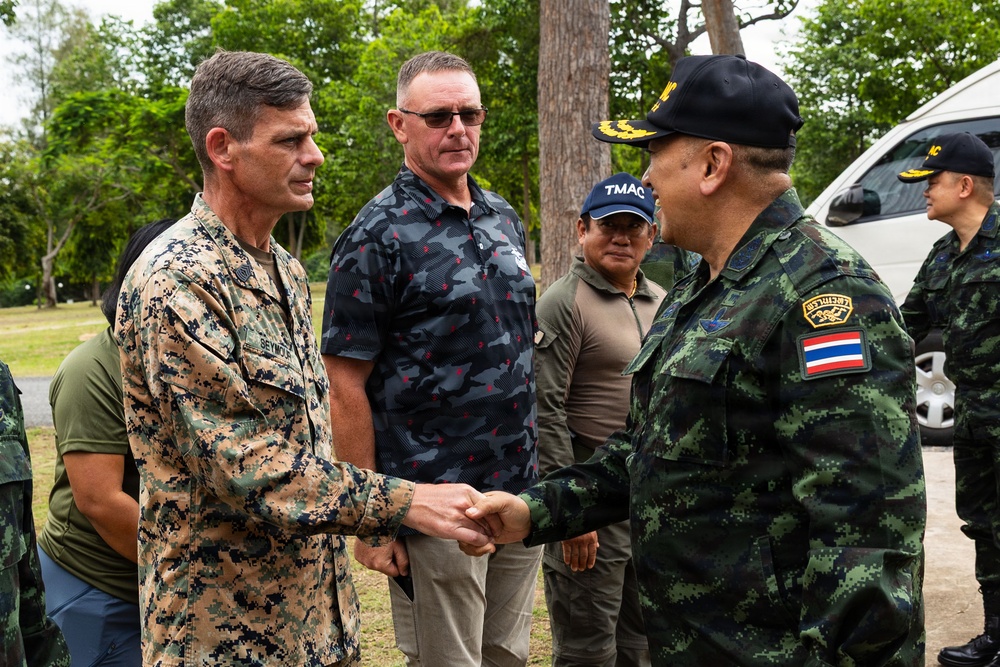 A formal military greeting is taking place between a U.S. Marine Corps officer and a Thai military officer, who are shaking hands. Several other individuals, including civilians and military personnel, are present in the background. The setting appears to be an outdoor area with trees and a vehicle visible, suggesting a field visit or joint military exercise.
