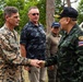 A formal military greeting is taking place between a U.S. Marine Corps officer and a Thai military officer, who are shaking hands. Several other individuals, including civilians and military personnel, are present in the background. The setting appears to be an outdoor area with trees and a vehicle visible, suggesting a field visit or joint military exercise.