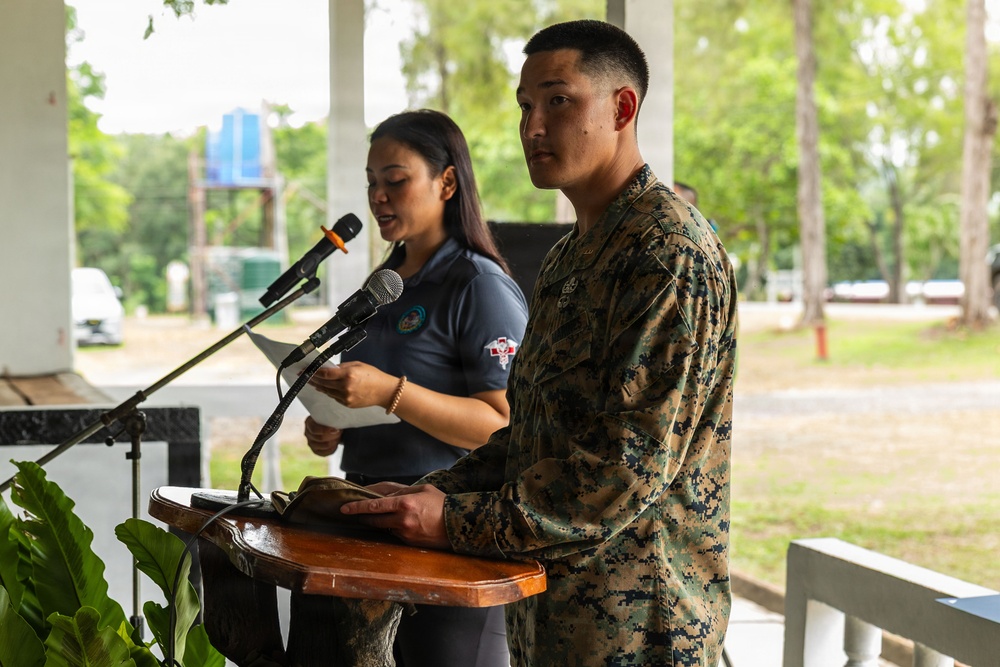 A U.S. Marine in woodland MARPAT camouflage uniform stands at a wooden podium during what appears to be an official ceremony or event. Behind him, a civilian woman in a dark blue polo shirt speaks into a separate microphone while holding papers, suggesting she may be serving as a translator or co-presenter. The setting appears to be an outdoor covered pavilion or veranda with green trees visible in the background.
