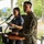 U.S. Marines and Thailand Mine Action Center participate in Deployed for Training Bilateral EOD Exercise A U.S. Marine in woodland MARPAT camouflage uniform stands at a wooden podium during what appears to be an official ceremony or event. Behind him, a civilian woman in a dark blue polo shirt speaks into a separate microphone while holding papers, suggesting she may be serving as a translator or co-presenter. The setting appears to be an outdoor covered pavilion or veranda with green trees visible in the background.