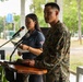 A U.S. Marine in woodland MARPAT camouflage uniform stands at a wooden podium during what appears to be an official ceremony or event. Behind him, a civilian woman in a dark blue polo shirt speaks into a separate microphone while holding papers, suggesting she may be serving as a translator or co-presenter. The setting appears to be an outdoor covered pavilion or veranda with green trees visible in the background.