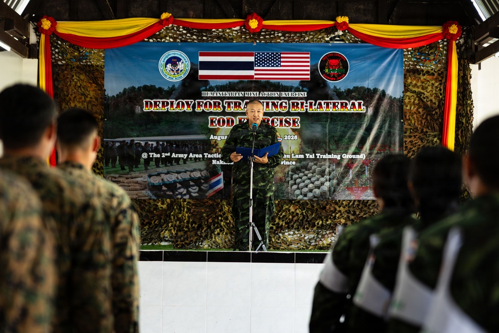 A military ceremony or briefing is taking place at a US-Thailand bilateral EOD (Explosive Ordnance Disposal) exercise, specifically a 'Humanitarian Mine Action Exercise - Deploy for Training Bi-Lateral EOD Exercise' held in August 2026 at the 2nd Army Area Tactical Training Ground (Lam Yai Training Ground) in Nakhon province, Thailand. A Thai military officer in camouflage uniform is speaking at a podium before an audience of soldiers in mixed US Marine and Thai military uniforms. The backdrop features Thai and American flags, unit insignia, and camouflage netting decorated with floral arrangements.