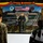 U.S. Marines and Thailand Mine Action Center participate in Deployed for Training Bilateral EOD Exercise A military ceremony or briefing is taking place at a US-Thailand bilateral EOD (Explosive Ordnance Disposal) exercise, specifically a 'Humanitarian Mine Action Exercise - Deploy for Training Bi-Lateral EOD Exercise' held in August 2026 at the 2nd Army Area Tactical Training Ground (Lam Yai Training Ground) in Nakhon province, Thailand. A Thai military officer in camouflage uniform is speaking at a podium before an audience of soldiers in mixed US Marine and Thai military uniforms. The backdrop features Thai and American flags, unit insignia, and camouflage netting decorated with floral arrangements.