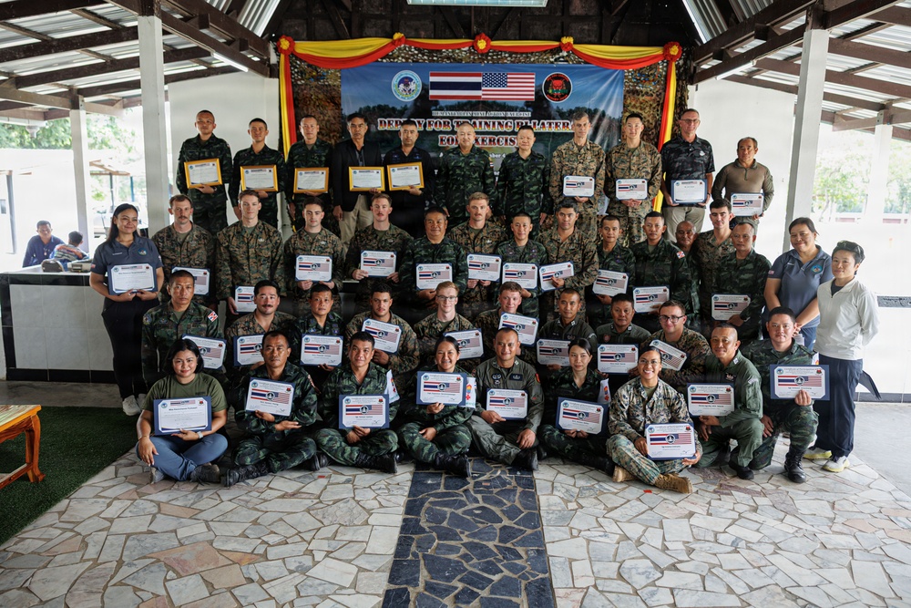 A large group of military personnel from Thailand and the United States pose together for a group photo holding certificates at what appears to be a bilateral training exercise completion ceremony. The backdrop displays a banner reading 'DEPLOY FOR TRAINING BILATERAL EOD EXERCISE' along with Thai and American flags and unit insignia. Participants are arranged in multiple rows under an open-air pavilion.