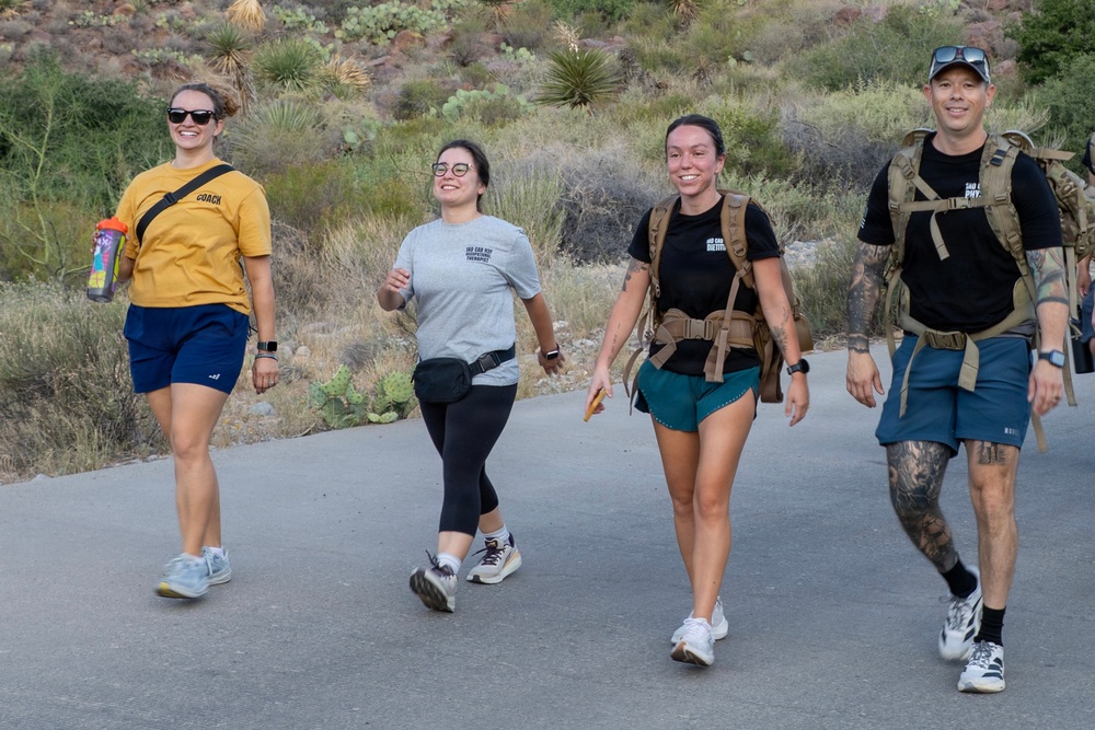 Four people are walking together on a paved path in what appears to be a desert or arid landscape, likely on a hiking trail. They are smiling and appear to be in good spirits, with two of them wearing tactical-style backpacks and one wearing a yellow shirt labeled 'COACH'. The background features desert vegetation including yucca plants and cacti, suggesting a southwestern U.S. location.