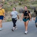 Four people are walking together on a paved path in what appears to be a desert or arid landscape, likely on a hiking trail. They are smiling and appear to be in good spirits, with two of them wearing tactical-style backpacks and one wearing a yellow shirt labeled 'COACH'. The background features desert vegetation including yucca plants and cacti, suggesting a southwestern U.S. location.