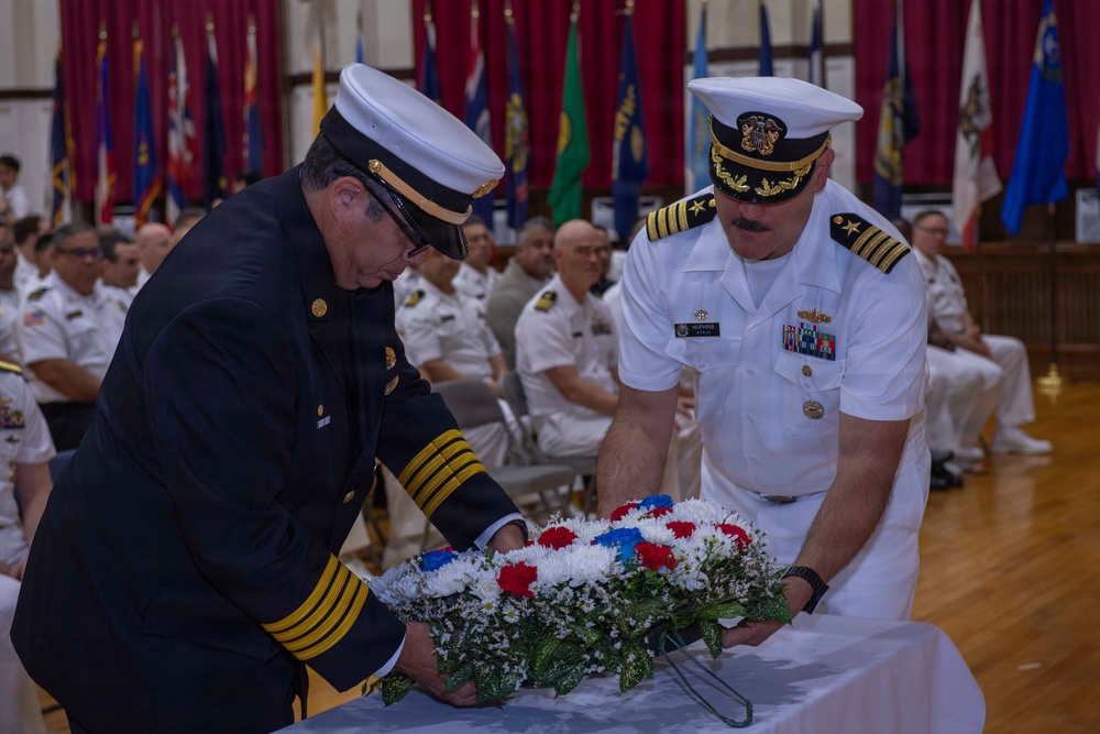 Two uniformed officials — one in a dark navy/fire department dress uniform and one in a white U.S. Navy officer's uniform — are jointly placing a red, white, and blue floral wreath on a table during a formal memorial or commemorative ceremony. The event is taking place in a large hall decorated with multiple military and state flags, with rows of seated uniformed personnel visible in the background. The solemn nature of the wreath-laying suggests this is a memorial service or official ceremony.