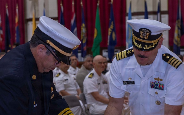 Two uniformed officials — one in a dark navy/fire department dress uniform and one in a white U.S. Navy officer's uniform — are jointly placing a red, white, and blue floral wreath on a table during a formal memorial or commemorative ceremony. The event is taking place in a large hall decorated with multiple military and state flags, with rows of seated uniformed personnel visible in the background. The solemn nature of the wreath-laying suggests this is a memorial service or official ceremony.
