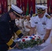 Two uniformed officials — one in a dark navy/fire department dress uniform and one in a white U.S. Navy officer's uniform — are jointly placing a red, white, and blue floral wreath on a table during a formal memorial or commemorative ceremony. The event is taking place in a large hall decorated with multiple military and state flags, with rows of seated uniformed personnel visible in the background. The solemn nature of the wreath-laying suggests this is a memorial service or official ceremony.