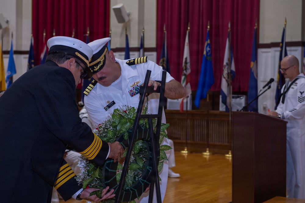 A formal military memorial ceremony is taking place in what appears to be a ceremonial hall. Two naval officers in dress uniforms are placing or adjusting a floral wreath on a stand, while a third sailor stands at a podium with a microphone. Multiple state and military flags are displayed in the background against red curtains.