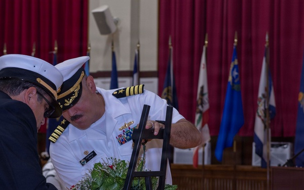 A formal military memorial ceremony is taking place in what appears to be a ceremonial hall. Two naval officers in dress uniforms are placing or adjusting a floral wreath on a stand, while a third sailor stands at a podium with a microphone. Multiple state and military flags are displayed in the background against red curtains.