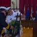 A formal military memorial ceremony is taking place in what appears to be a ceremonial hall. Two naval officers in dress uniforms are placing or adjusting a floral wreath on a stand, while a third sailor stands at a podium with a microphone. Multiple state and military flags are displayed in the background against red curtains.