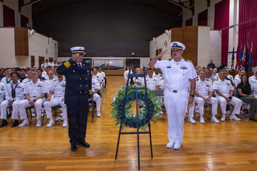 A formal military memorial ceremony is taking place in a large auditorium or hall, with two senior officers — one in a dark navy/fire department dress uniform and one in a white naval officer uniform — rendering salutes beside a wreath on a stand. A large audience of uniformed military personnel is seated in rows behind them. The setting and wreath suggest a commemorative or memorial service.