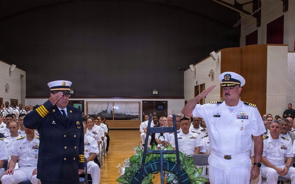 A formal military memorial ceremony is taking place in a large auditorium or hall, with two senior officers — one in a dark navy/fire department dress uniform and one in a white naval officer uniform — rendering salutes beside a wreath on a stand. A large audience of uniformed military personnel is seated in rows behind them. The setting and wreath suggest a commemorative or memorial service.