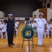 A formal military memorial ceremony is taking place in a large auditorium or hall, with two senior officers — one in a dark navy/fire department dress uniform and one in a white naval officer uniform — rendering salutes beside a wreath on a stand. A large audience of uniformed military personnel is seated in rows behind them. The setting and wreath suggest a commemorative or memorial service.