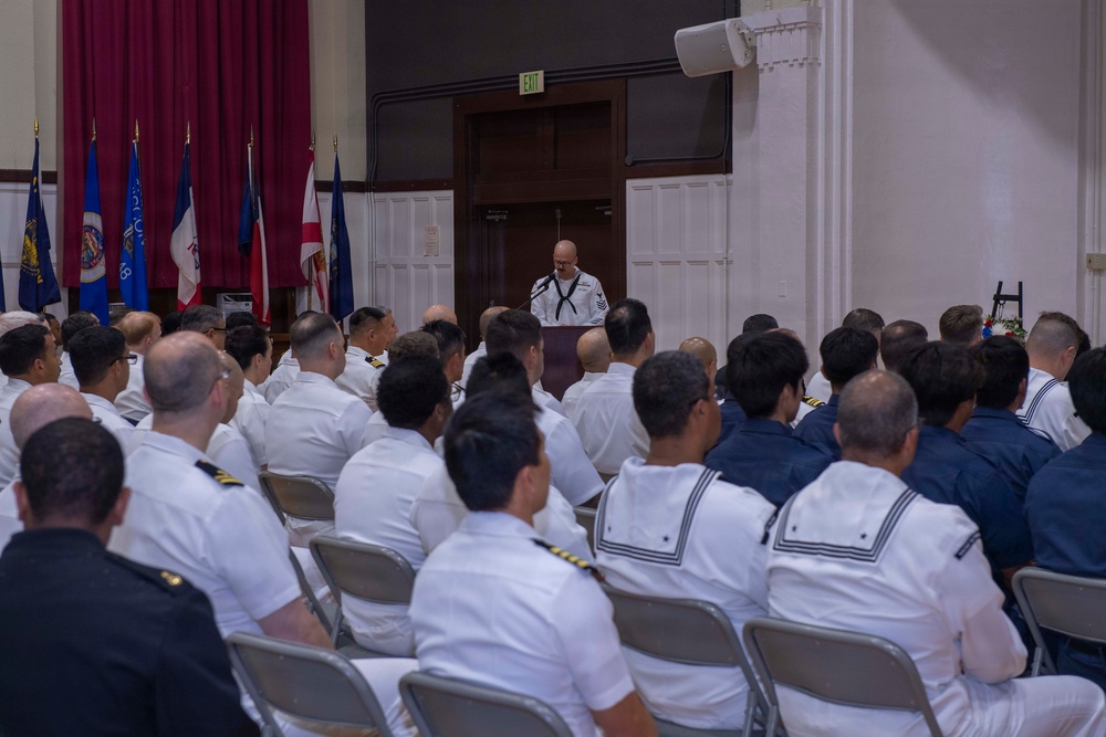 A military ceremony or formal gathering is taking place in an indoor hall, with a large audience of naval personnel seated in rows of folding chairs. A speaker in white Navy uniform stands at a podium addressing the assembled crowd. Multiple national and organizational flags are displayed on the left side of the room near a stage area.