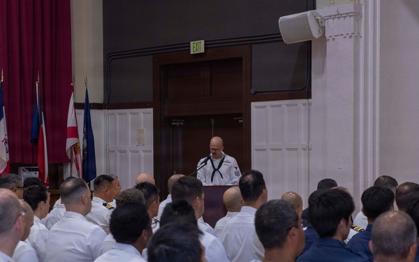 A military ceremony or formal gathering is taking place in an indoor hall, with a large audience of naval personnel seated in rows of folding chairs. A speaker in white Navy uniform stands at a podium addressing the assembled crowd. Multiple national and organizational flags are displayed on the left side of the room near a stage area.