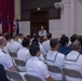 A military ceremony or formal gathering is taking place in an indoor hall, with a large audience of naval personnel seated in rows of folding chairs. A speaker in white Navy uniform stands at a podium addressing the assembled crowd. Multiple national and organizational flags are displayed on the left side of the room near a stage area.