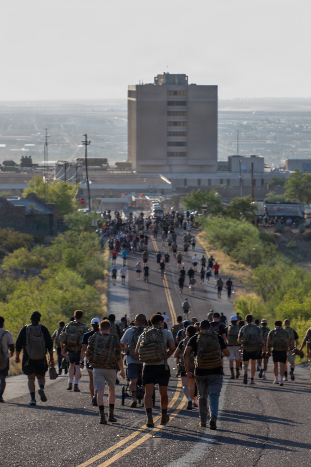 A large group of people, many wearing military-style tactical backpacks and boots, march down a two-lane road with double yellow center lines. The procession stretches far into the distance toward a mid-rise building in what appears to be a desert or semi-arid urban environment. The scene is consistent with a memorial ruck march or organized military/fitness event.