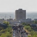 A large group of people, many wearing military-style tactical backpacks and boots, march down a two-lane road with double yellow center lines. The procession stretches far into the distance toward a mid-rise building in what appears to be a desert or semi-arid urban environment. The scene is consistent with a memorial ruck march or organized military/fitness event.