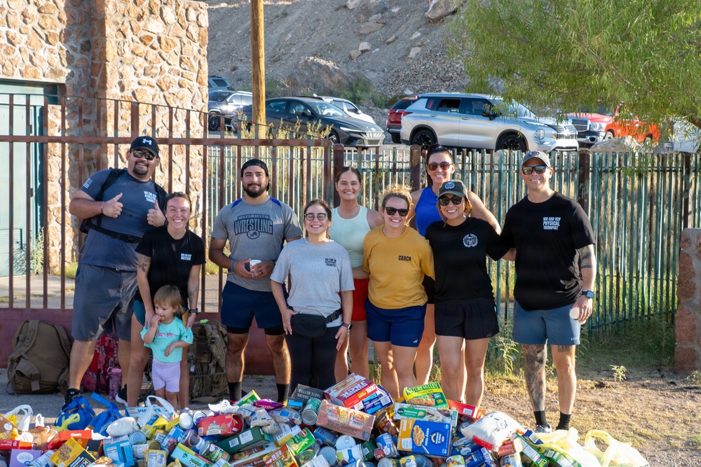 A group of approximately nine adults and one toddler pose together outdoors in front of a large collection of donated food items including canned goods, granola bars, and packaged foods. The setting appears to be near a stone building with an iron fence, with a parking lot and rocky hillside visible in the background. Several individuals are wearing matching black t-shirts suggesting organizational affiliation, and one person wears a yellow shirt labeled 'COACH'.