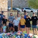 A group of approximately nine adults and one toddler pose together outdoors in front of a large collection of donated food items including canned goods, granola bars, and packaged foods. The setting appears to be near a stone building with an iron fence, with a parking lot and rocky hillside visible in the background. Several individuals are wearing matching black t-shirts suggesting organizational affiliation, and one person wears a yellow shirt labeled 'COACH'.