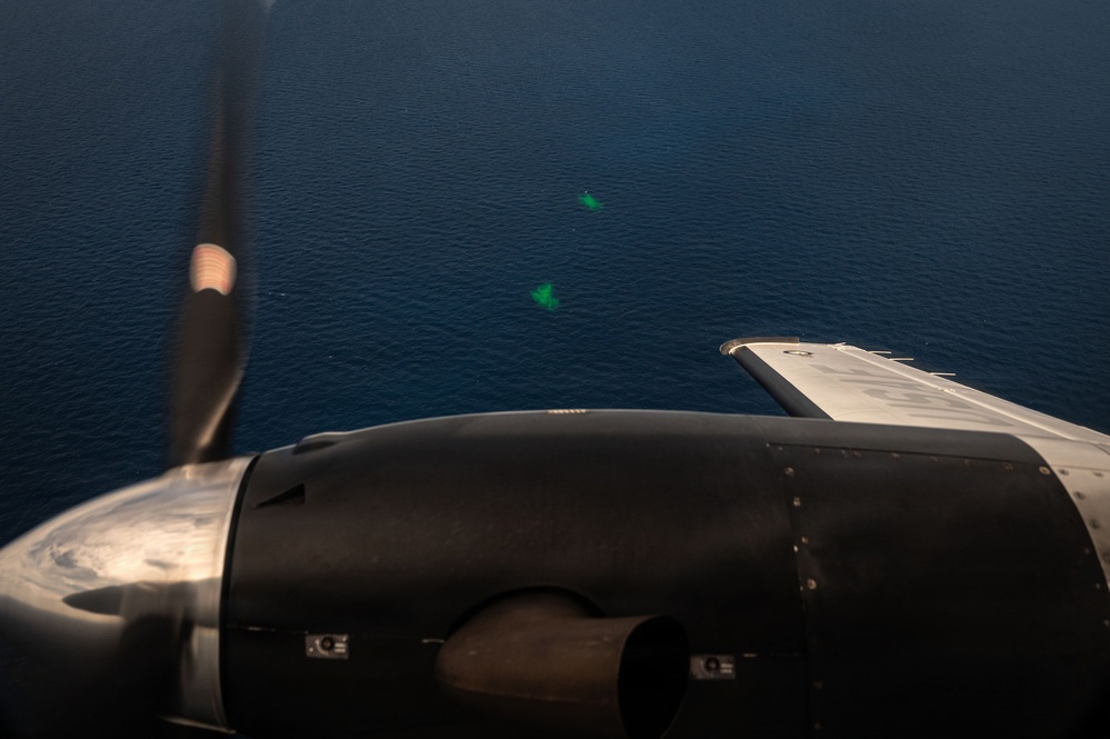 An aerial photograph taken from a propeller-driven aircraft flying over open ocean. Two small bright green markers or dye patches are visible on the water surface below, consistent with maritime search and rescue or military marking operations. The image is shot through or past the aircraft's engine nacelle and spinning propeller.