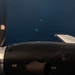 An aerial photograph taken from a propeller-driven aircraft flying over open ocean. Two small bright green markers or dye patches are visible on the water surface below, consistent with maritime search and rescue or military marking operations. The image is shot through or past the aircraft's engine nacelle and spinning propeller.
