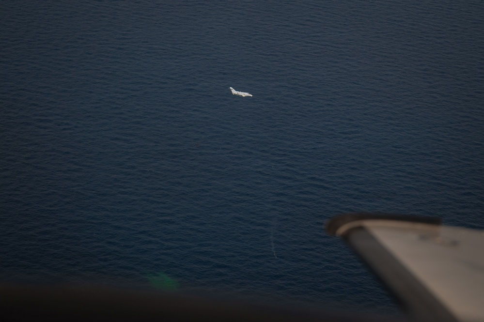 An aerial photograph taken from inside an aircraft looking down at a large body of dark blue water. A second aircraft is visible at a lower altitude in the middle distance, appearing small against the ocean surface. The wing or engine nacelle of the photographing aircraft is partially visible in the lower right corner of the frame.