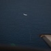 An aerial photograph taken from inside an aircraft looking down at a large body of dark blue water. A second aircraft is visible at a lower altitude in the middle distance, appearing small against the ocean surface. The wing or engine nacelle of the photographing aircraft is partially visible in the lower right corner of the frame.