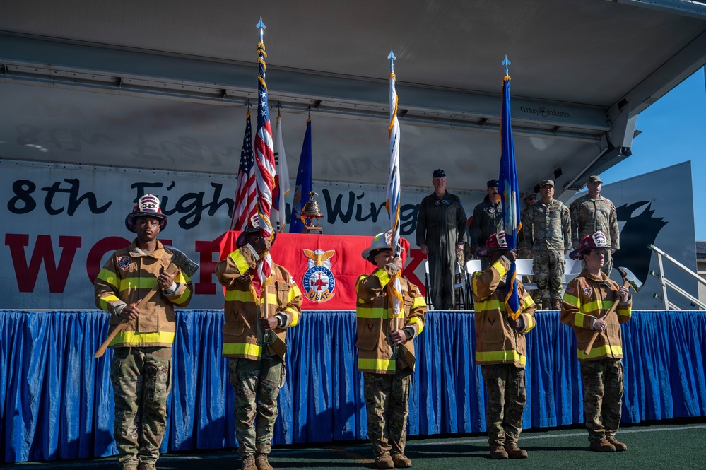 A formal military ceremony is taking place at the 8th Fighter Wing, likely at Kunsan Air Base. A color guard composed of personnel wearing firefighter turnout gear over military uniforms is presenting flags, including the U.S. flag and USAF Fire Protection flag. Military officers and personnel stand on a decorated stage behind them.