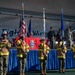 A formal military ceremony is taking place at the 8th Fighter Wing, likely at Kunsan Air Base. A color guard composed of personnel wearing firefighter turnout gear over military uniforms is presenting flags, including the U.S. flag and USAF Fire Protection flag. Military officers and personnel stand on a decorated stage behind them.
