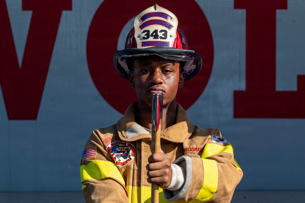 A young firefighter in full turnout gear poses confidently while holding a tool, likely a halligan bar or axe handle. They are wearing a white and red helmet marked '343' with 'Wolfpack Fire/Rescue' and 'Fire Guard' text. The subject stands in front of a wall with large red letters partially spelling 'VOL', suggesting a volunteer fire department context.