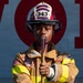 A young firefighter in full turnout gear poses confidently while holding a tool, likely a halligan bar or axe handle. They are wearing a white and red helmet marked '343' with 'Wolfpack Fire/Rescue' and 'Fire Guard' text. The subject stands in front of a wall with large red letters partially spelling 'VOL', suggesting a volunteer fire department context.