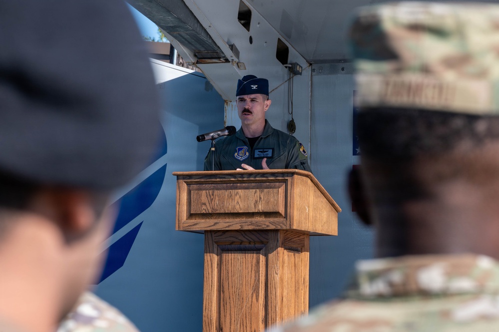 A U.S. Air Force officer wearing a flight suit and colonel's rank insignia (silver eagle on cap) is speaking at a wooden podium with a microphone. The setting appears to be an outdoor military ceremony or event, with uniformed personnel visible in the foreground as audience members. The speaker has a mustache and wears unit patches on his flight suit.