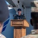 A U.S. Air Force officer wearing a flight suit and colonel's rank insignia (silver eagle on cap) is speaking at a wooden podium with a microphone. The setting appears to be an outdoor military ceremony or event, with uniformed personnel visible in the foreground as audience members. The speaker has a mustache and wears unit patches on his flight suit.