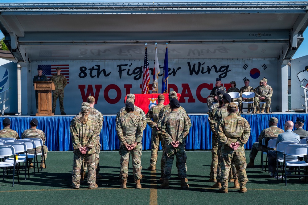 A formal military ceremony is taking place at the 8th Fighter Wing 'Wolfpack' unit, with personnel standing at attention facing a decorated stage. The event appears to be an official function such as a change of command, award ceremony, or unit activation, held outdoors on what appears to be a sports court. Flags of the United States, U.S. Air Force, and South Korea are prominently displayed on stage, indicating this is likely at Kunsan Air Base, South Korea.