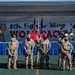 A formal military ceremony is taking place at the 8th Fighter Wing 'Wolfpack' unit, with personnel standing at attention facing a decorated stage. The event appears to be an official function such as a change of command, award ceremony, or unit activation, held outdoors on what appears to be a sports court. Flags of the United States, U.S. Air Force, and South Korea are prominently displayed on stage, indicating this is likely at Kunsan Air Base, South Korea.