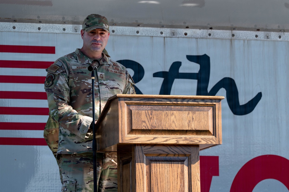 A U.S. Air Force service member identified by name tape as 'AGUIAR' stands at a wooden podium delivering a speech at what appears to be a military ceremony or event. The individual is wearing OCP (Operational Camouflage Pattern) uniform with U.S. Air Force branch tape and a senior enlisted rank insignia visible on the chest. An American flag and partial text reading '8th' are visible in the background.