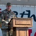 A U.S. Air Force service member identified by name tape as 'AGUIAR' stands at a wooden podium delivering a speech at what appears to be a military ceremony or event. The individual is wearing OCP (Operational Camouflage Pattern) uniform with U.S. Air Force branch tape and a senior enlisted rank insignia visible on the chest. An American flag and partial text reading '8th' are visible in the background.
